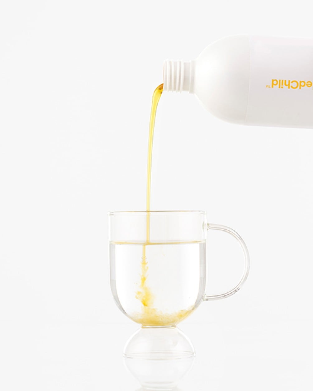 A white bottle pouring golden pineapple liquid collagen into a clear glass mug of water against a white background.